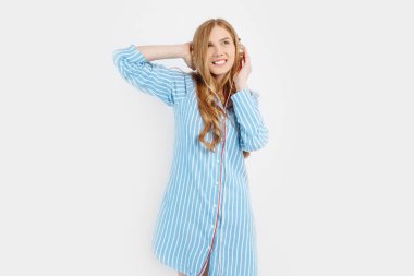 Mutlu, düşünceli, çekici, pijamalı, eğlenceli ve dans eden beyaz bir arka planda kulaklıkla müzik dinleyen genç kız.