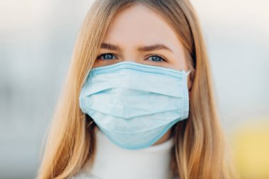 Binanın arka planındaki genç bir kız, koronavirüs hastalığının yayılmasını önleyen bir maske takıyor. Yüzünde cerrahi maske olan genç bir kadının SARS-cov-2 'ye yakın çekimi..