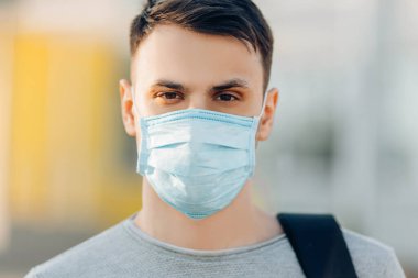 Açık hava binasının arka planında, Coronavirus hastalığının yayılmasını önleyen tıbbi bir maske takan genç bir adam. Yüzünde cerrahi maske olan bir adamın yakın çekimi.