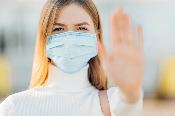 Yüzünde koruyucu steril tıbbi maske takmış bir kadın açık havada kameraya bakıyor, bir avuç içi, bir el, bir dur işareti, işaretsiz bir durak gösteriyor. Çin virüs salgını konsepti.