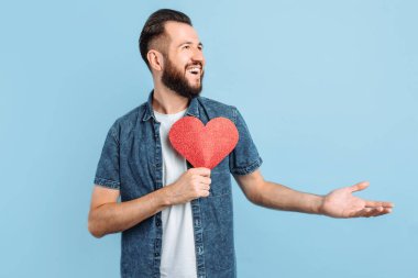 Aşk ve Sevgililer Günü 'nde sakallı çekici bir adam kırmızı kağıt bir kalp taşıyor, gülümsüyor, mavi bir arka planda. Sevgililer Günü. Kırmızı kalp.