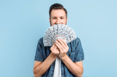 Neşeli, heyecanlı genç bir adam mavi bir arka planda poz veriyor, elinde bir dolar banknotları olan bir nakit hayranı var. İnsanların yaşam tarzı kavramı.