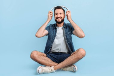 Günlük giysiler içinde, kulaklıkla müzik dinleyen, mavi arka planda oturan mutlu, genç yakışıklı bir adam.