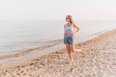 Güneş gözlüklü çekici genç kız, kumsalda geziniyor güneşli havanın tadını çıkarıyor..