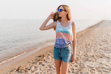 Güneş gözlüklü çekici genç kız, kumsalda geziniyor güneşli havanın tadını çıkarıyor..