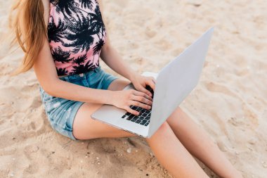 Kız kumsalda kumda oturan bir dizüstü bilgisayarla çalışıyor.