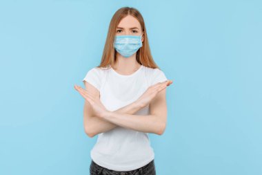 Koruyucu tıbbi maske takan genç bir kadın çapraz kollu ve hava kirliliğiyle durdurma hareketi gösteriyor. Çin koronavirüs salgınına karşı izole edilmiş mavi arka planda koruma.