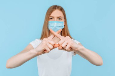 Koruyucu tıbbi maske takan genç bir kadın, parmaklarını çapraz yapıp, hava kirliliğini durdurma hareketi gösteriyor. Çin koronavirüs salgınına karşı izole edilmiş mavi arka planda koruma.