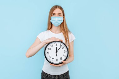 Yüzünde Coronavirus 'a karşı koruyucu bir maske takan genç bir kadın izole edilmiş mavi bir arka planda duvar saati tutuyor. Hava kirliliği, virüs, koronavirüs