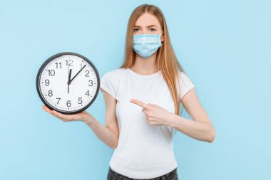 Yüzünde Coronavirus 'a karşı koruyucu bir maske takan genç bir kadın izole edilmiş mavi bir arka planda duvar saati tutuyor. Hava kirliliği, virüs, koronavirüs