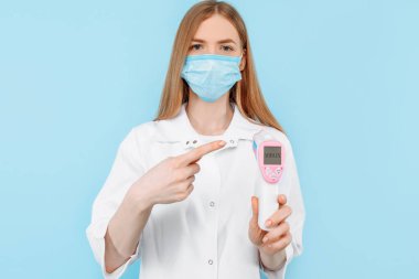 Tıbbi maske takan ve beyaz önlük giyen bir kadın doktor, hastanın vücut sıcaklığını mavi bir arka planla karşılaştırmak için temas edilmeyen kızılötesi bir alın termometresi kullanır..