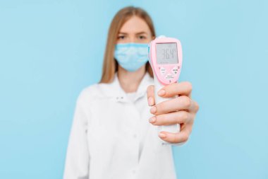 Tıbbi maske takan genç bir bayan doktor ve beyaz laboratuvar önlüğü normal vücut sıcaklığı 36.4 derece olan temassız termometre ekranını gösteriyor. Tıbbi bakım kavramı, virüs salgını