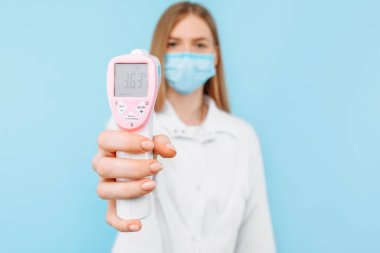 Tıbbi maske takan genç bir bayan doktor ve beyaz laboratuvar önlüğü normal vücut sıcaklığı 36.3 derece olan temassız termometre ekranını gösteriyor. Tıbbi bakım kavramı, virüs salgını