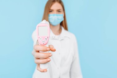 Tıbbi maske takan genç bir bayan doktor ve beyaz laboratuvar önlüğü normal vücut sıcaklığı 36.4 derece olan temassız termometre ekranını gösteriyor. Tıbbi bakım kavramı, virüs salgını