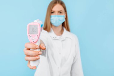 Tıbbi maske takan genç bir bayan doktor ve beyaz laboratuvar önlüğü normal vücut sıcaklığı 36.3 derece olan temassız termometre ekranını gösteriyor. Tıbbi bakım kavramı, virüs salgını