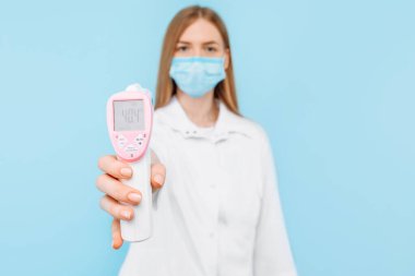 Tıbbi maske takan ve beyaz laboratuvar önlüğü giyen genç bir kadın doktor, 40 derece yüksek vücut ısısına sahip temas edilmeyen termometre ekranını gösteriyor. Tıbbi bakım kavramı