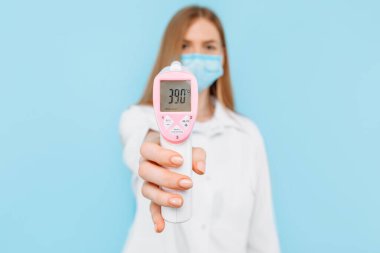 Tıbbi maske takan ve beyaz laboratuvar önlüğü giyen genç bir kadın doktor, 39.0 derece yüksek vücut ısısına sahip temas edilmeyen termometre ekranını gösteriyor. Tıbbi bakım kavramı
