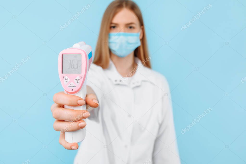 A young female doctor wearing a medical mask and a white lab coat shows the screen of a non-contact thermometer with a normal body temperature of 36.3 degrees. Concept of medical care, virus outbreak