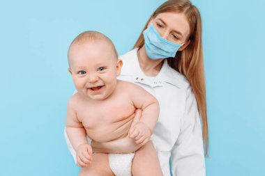 Tıp, sağlık ve pediatri konsepti, dijital kızılötesi termometresi olan kadın bir çocuk doktoru, bebeğin vücut ısısını izole edilmiş mavi bir arka planda ölçer.
