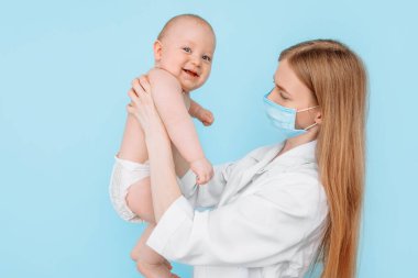 Tıp, sağlık ve pediatri konsepti, dijital kızılötesi termometresi olan kadın bir çocuk doktoru, bebeğin vücut ısısını izole edilmiş mavi bir arka planda ölçer.