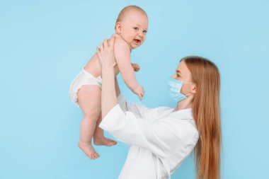Tıp, sağlık ve pediatri konsepti, dijital kızılötesi termometresi olan kadın bir çocuk doktoru, bebeğin vücut ısısını izole edilmiş mavi bir arka planda ölçer.