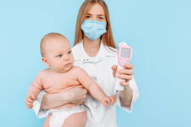 Tıp, Sağlık ve Pediatri konsepti. İzole edilmiş mavi arka planda bebeğin alnının sıcaklığını ölçen dijital kızılötesi termometresi olan kadın bir çocuk doktoru.