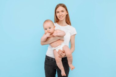 Genç, narin, mutlu bir anne, yeni doğmuş bebeğine sarılıyor, gülümsüyor, izole bir mavi arka planda. Annelik kavramı, bebek bezi.