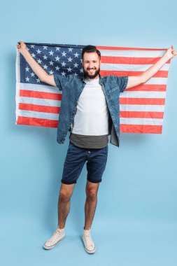 Bağımsızlık Günü kutlaması. Yıldızlar ve çizgiler. Amerika Birleşik Devletleri bayrağına sahip genç bir adam, izole edilmiş mavi bir arka planda, ülkesinin bir vatansever gibi mutlu görünüyor.