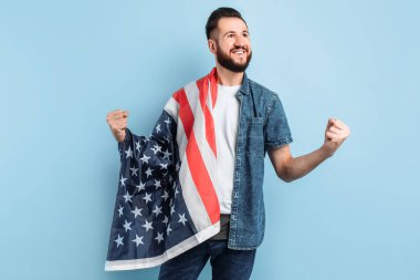 Sakallı mutlu bir gencin portresi, günlük giysiler içinde, Amerikan bayrağını tutuyor ve mavi bir arka planda zafer ve başarı jesti sergiliyor.