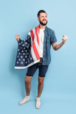 Sakallı mutlu bir gencin portresi, günlük giysiler içinde, Amerikan bayrağını tutuyor ve mavi bir arka planda zafer ve başarı jesti sergiliyor.