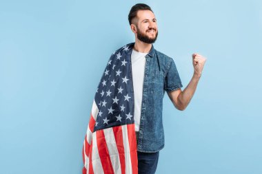 Sakallı mutlu bir gencin portresi, günlük giysiler içinde, Amerikan bayrağını tutuyor ve mavi bir arka planda zafer ve başarı jesti sergiliyor.