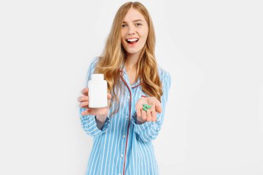 Pijamalı bir kız elinde beyaz plastik bir hap kavanozu, hastalıkların tıbbi tedavisi için bir konsept, beyaz bir arka planda beyaz bir hap kavanozunun yakın çekimi,