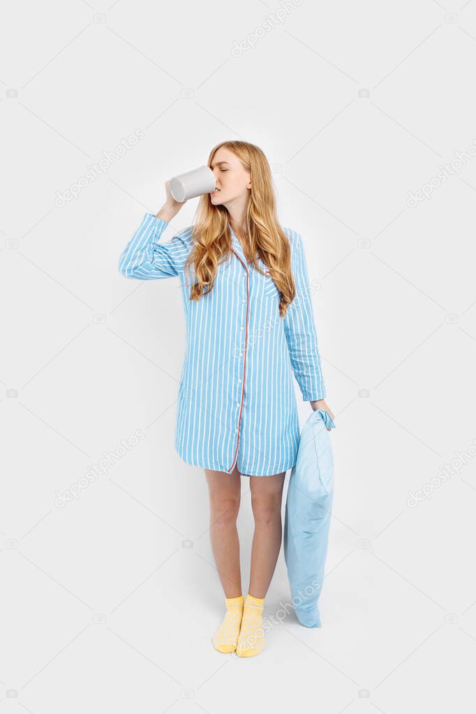 Beautiful girl in striped pajamas, with a pillow in her hands , with a Cup in her hands, drinking coffee or tea in the early morning, on a white background, isolated for text