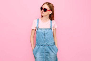 Güneş gözlüklü, pembe bir arka planda poz veren mutlu stil sahibi çekici genç kız.