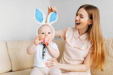 Anne ve bebek tavşan kulaklı, ellerinde paskalya yumurtaları, kanepede oturuyorlar, aileler ve çocuklar içeride oynuyorlar. Aile Paskalyayı kutluyor