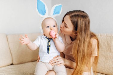 Anne ve bebek tavşan kulaklı, ellerinde paskalya yumurtaları, kanepede oturuyorlar, aileler ve çocuklar içeride oynuyorlar. Aile Paskalyayı kutluyor