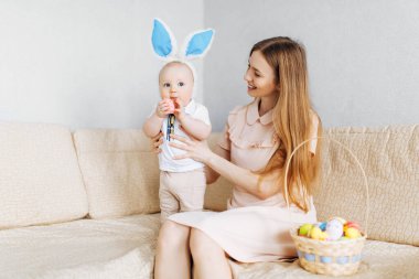 Tavşan kulaklı anne ve bebek sepetinde Paskalya yumurtaları ile kanepede oturuyor, ebeveynler ve çocuklar içeride oynuyor. Aile Paskalyayı kutlar, aile tatili.