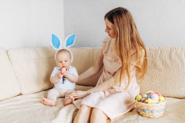 Tavşan kulaklı anne ve bebek sepetinde Paskalya yumurtaları ile kanepede oturuyor, ebeveynler ve çocuklar içeride oynuyor. Aile Paskalyayı kutlar, aile tatili.