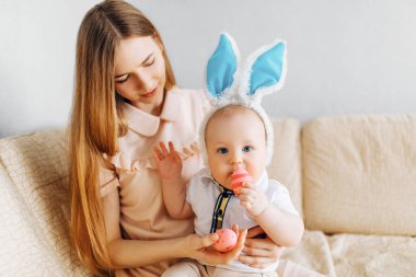 Anne ve bebek tavşan kulaklı, ellerinde paskalya yumurtaları, kanepede oturuyorlar, aileler ve çocuklar içeride oynuyorlar. Aile Paskalyayı kutluyor