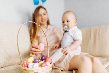 Tavşan kulaklı anne ve bebek sepetinde Paskalya yumurtaları ile kanepede oturuyor, ebeveynler ve çocuklar içeride oynuyor. Aile Paskalyayı kutlar, aile tatili.