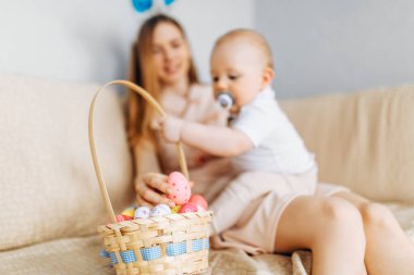 Tavşan kulaklı anne ve bebek sepetinde Paskalya yumurtaları ile kanepede oturuyor, ebeveynler ve çocuklar içeride oynuyor. Aile Paskalyayı kutlar, aile tatili.