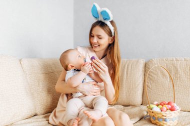 Tavşan kulaklı anne ve bebek sepetinde Paskalya yumurtaları ile kanepede oturuyor, ebeveynler ve çocuklar içeride oynuyor. Aile Paskalyayı kutlar, aile tatili.