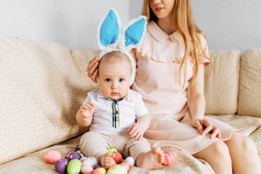 Mutlu Paskalyalar. Paskalya günü tavşan kulaklı bir anne ve küçük bir bebek, Paskalya yumurtası boyalı, eğlen, oyna ve sarıl, kanepede otur. Aile Paskalyayı kutluyor