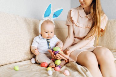Mutlu Paskalyalar. Paskalya günü tavşan kulaklı bir anne ve küçük bir bebek, Paskalya yumurtası boyalı, eğlen, oyna ve sarıl, kanepede otur. Aile Paskalyayı kutluyor