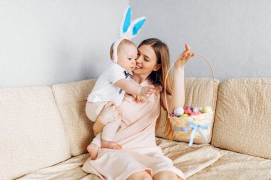 Mutlu Paskalyalar. Paskalya günü tavşan kulaklı bir anne ve küçük bir bebek, Paskalya yumurtası boyalı, eğlen, oyna ve sarıl, kanepede otur. Aile Paskalyayı kutluyor