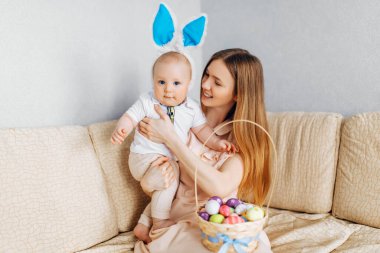 Mutlu Paskalyalar. Paskalya günü tavşan kulaklı bir anne ve küçük bir bebek, Paskalya yumurtası boyalı, eğlen, oyna ve sarıl, kanepede otur. Aile Paskalyayı kutluyor