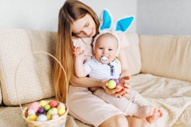 Mutlu Paskalyalar. Paskalya günü tavşan kulaklı bir anne ve küçük bir bebek, Paskalya yumurtası boyalı, eğlen, oyna ve sarıl, kanepede otur. Aile Paskalyayı kutluyor