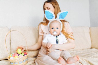 Mutlu Paskalyalar. Paskalya günü tavşan kulaklı bir anne ve küçük bir bebek, Paskalya yumurtası boyalı, eğlen, oyna ve sarıl, kanepede otur. Aile Paskalyayı kutluyor