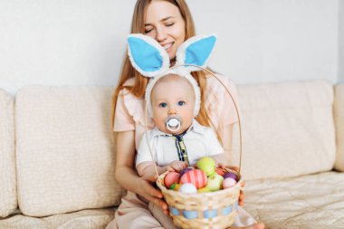 Mutlu Paskalyalar. Paskalya günü tavşan kulaklı bir anne ve küçük bir bebek, Paskalya yumurtası boyalı, eğlen, oyna ve sarıl, kanepede otur. Aile Paskalyayı kutluyor