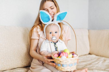 Mutlu Paskalyalar. Paskalya günü tavşan kulaklı bir anne ve küçük bir bebek, Paskalya yumurtası boyalı, eğlen, oyna ve sarıl, kanepede otur. Aile Paskalyayı kutluyor